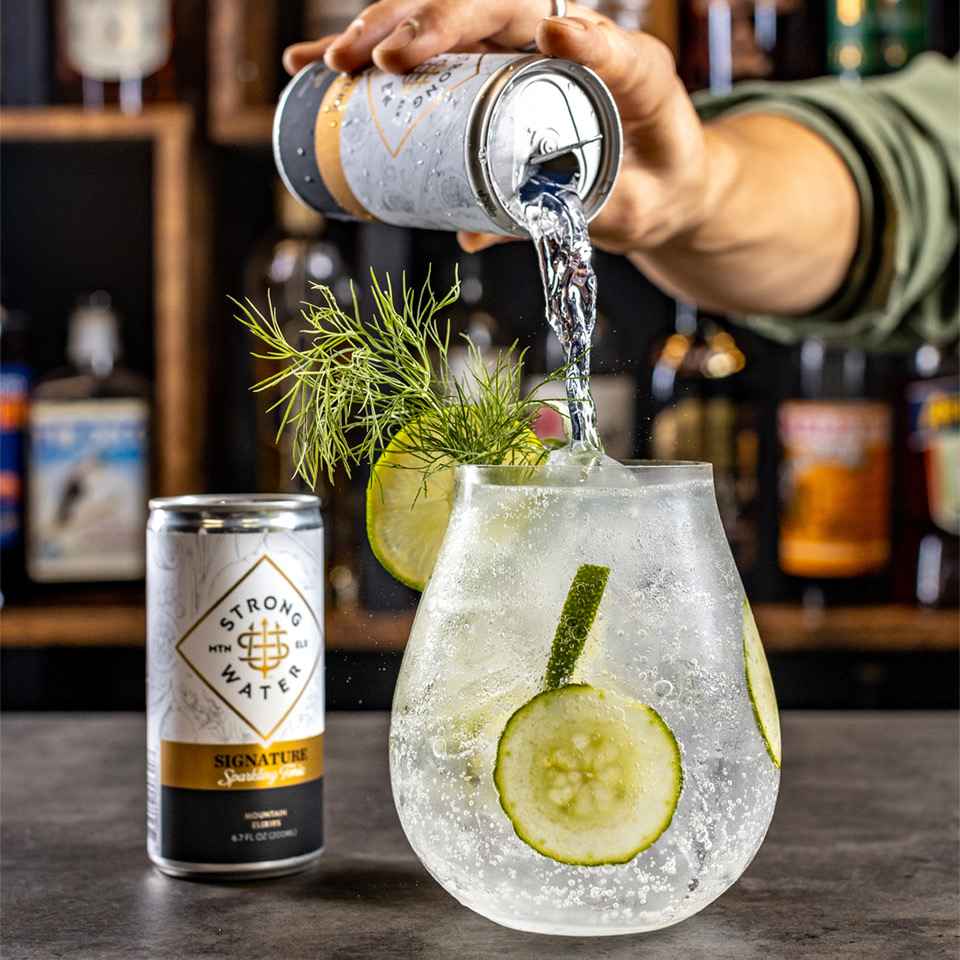 Hand pouring Strongwater mixer Signature Sparkling tonic into large cocktail glass garnished with cucumber, dill, and lemon.