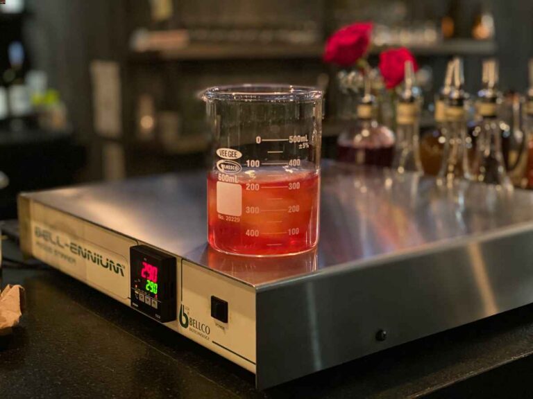 Lab beaker filled with ice and red cocktail sitting on a magnetic stir plate with bottles in background. Social in Ft. Collins, technology in restaurants.