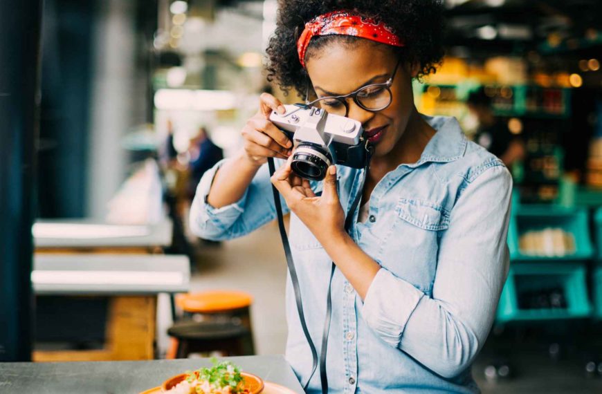 Black woman with natural hair using a film camera to photograph a plate of food.