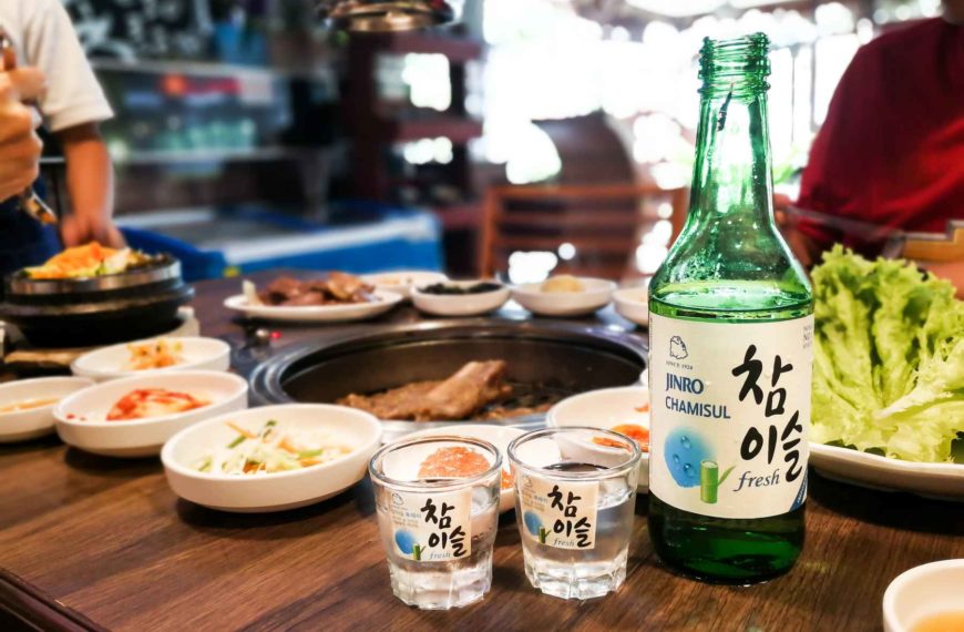 Bottle of soju and two shot glasses on a restaurant table next to a spread of Malaysian food.