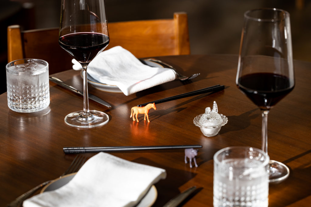 A table at Koko Ni restaurant in Denver set with plates, napkins, glasses of water and red wine, and chopsticks held by tiny toy horses. 