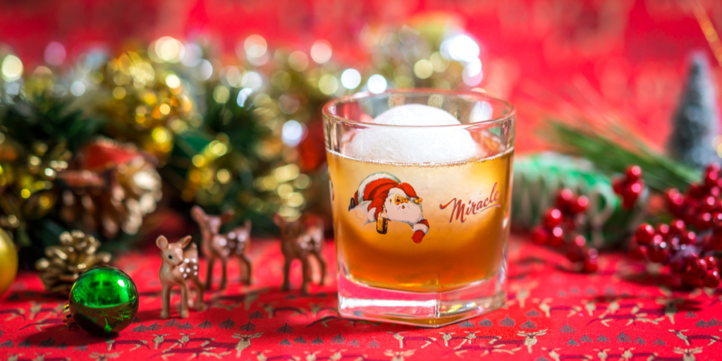 A christmas-themed old-fashioned cocktail in a small rocks glass on a red background with holiday decor