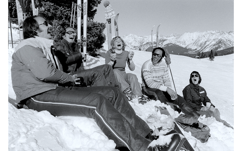 A vintage black-and-white photo of a group of people sitting in the snow mid-mountain at Vail in the 1970s