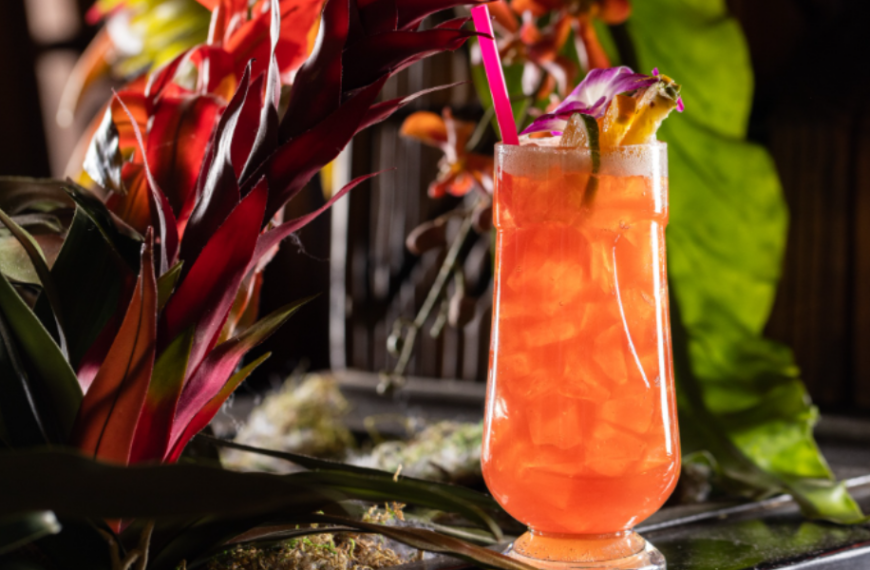 A vibrant orange-pink tropical cocktail from Adrift Tiki Bar in Denver is garnished with fresh fruit and a purple orchid. It has a neon pink straw and is set against a backdrop of tropical flora