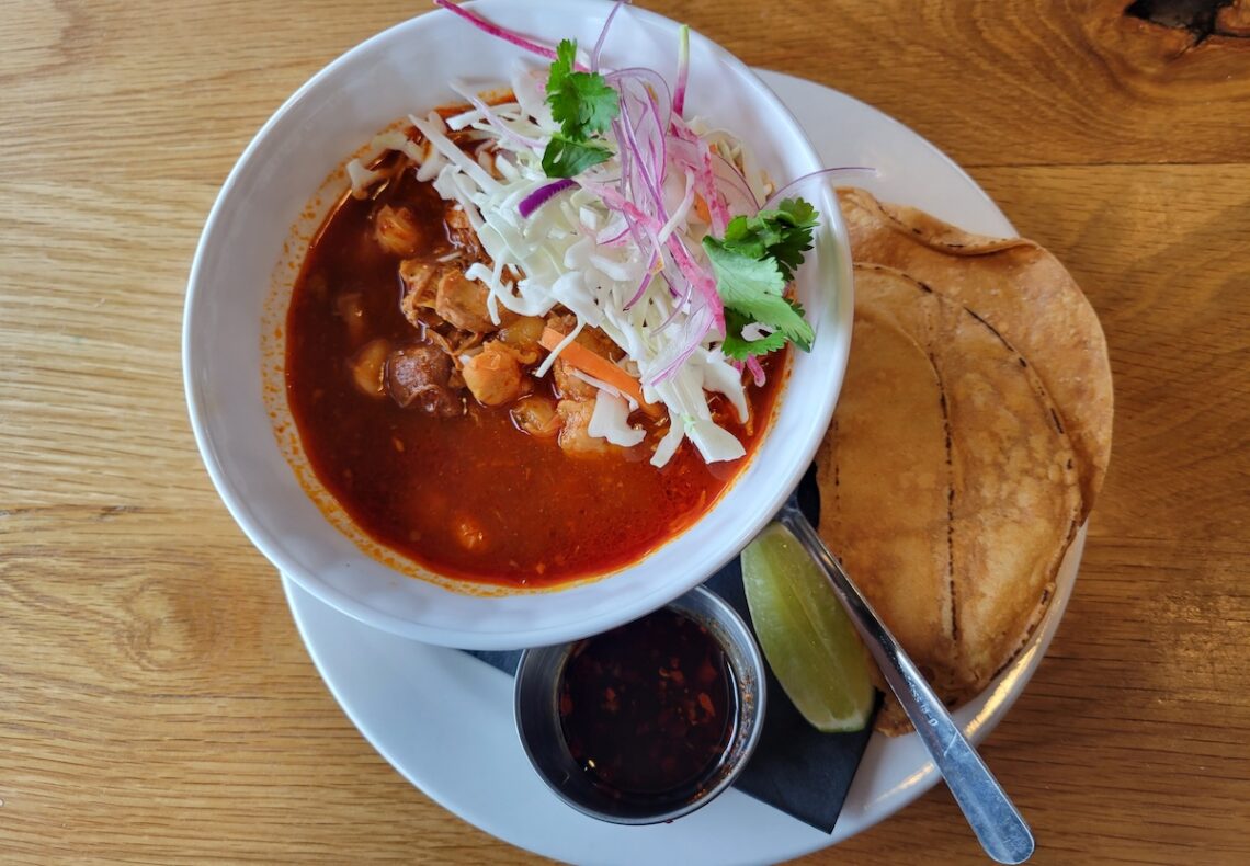 bowl of red pozole soup with tortilla on wood table