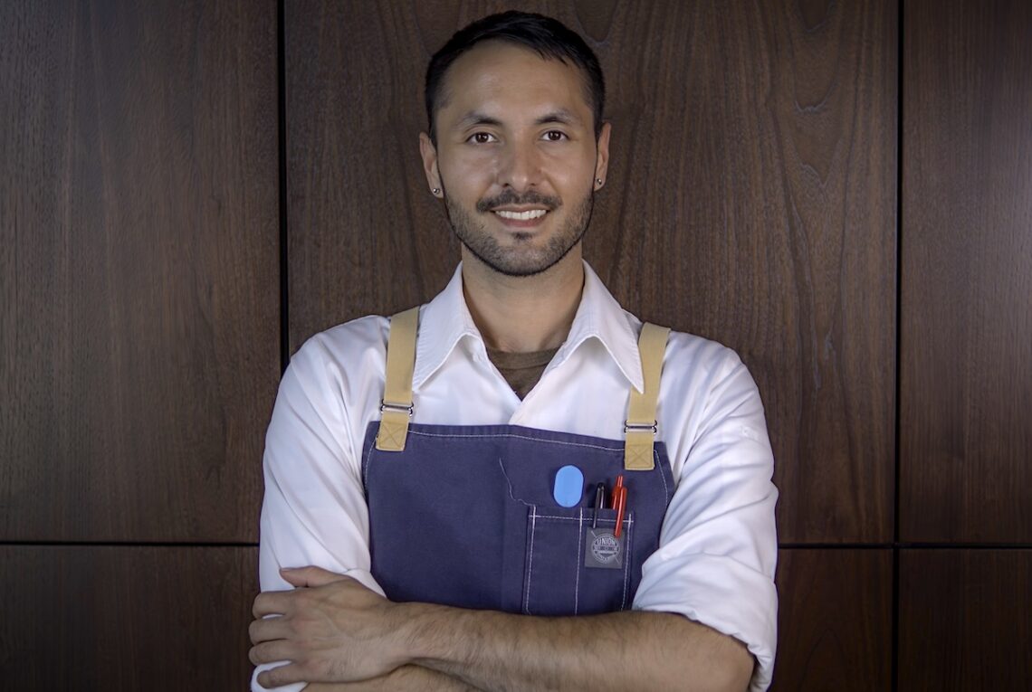 chef stands in front of wooded wall wearing blue apron