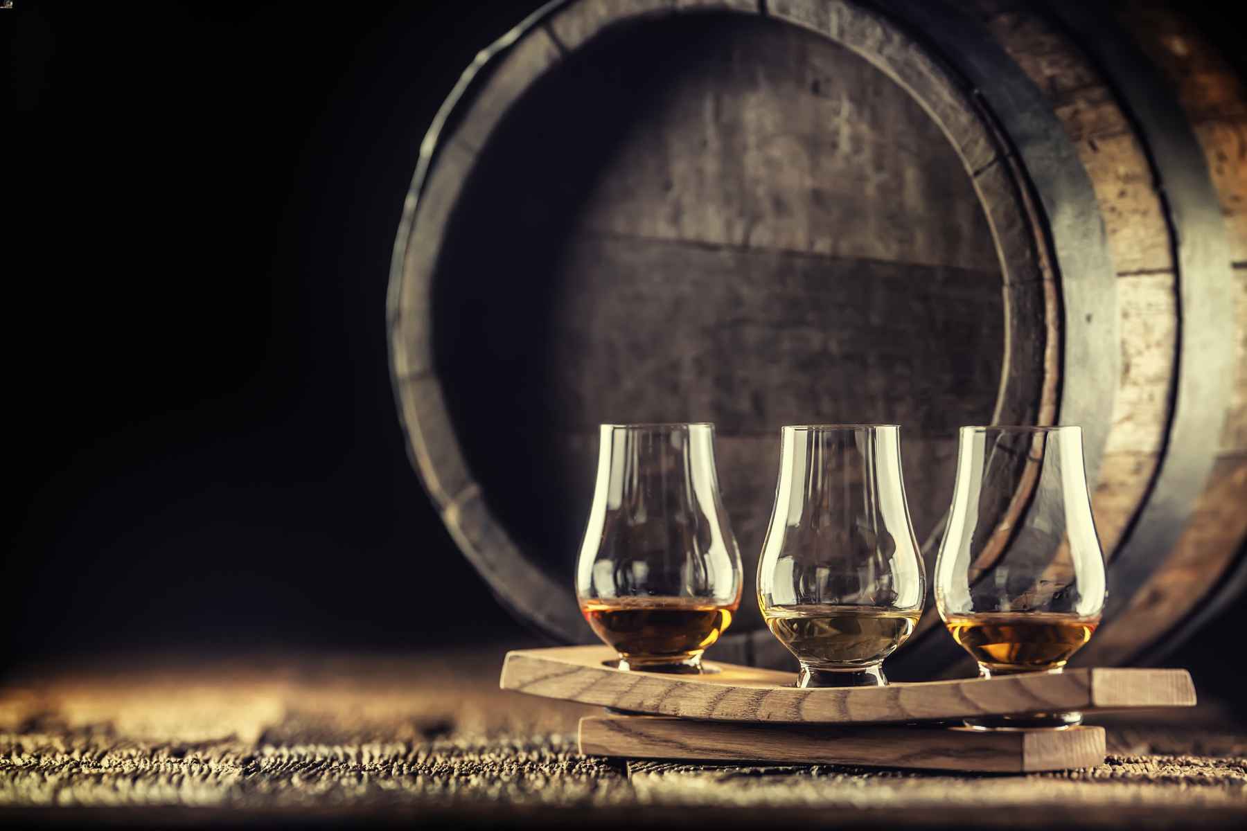 Three whiskey tasting glasses filled with whiskey in front of a barrel.
