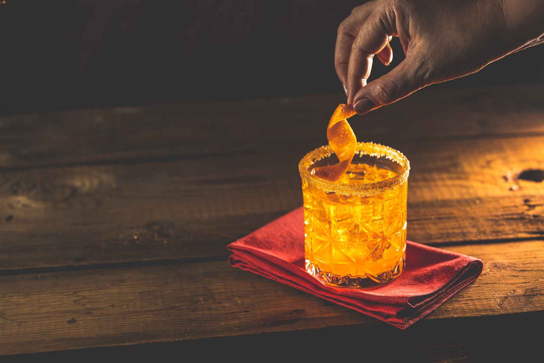Hand garnishing a cocktail with an orange with an orange twist.