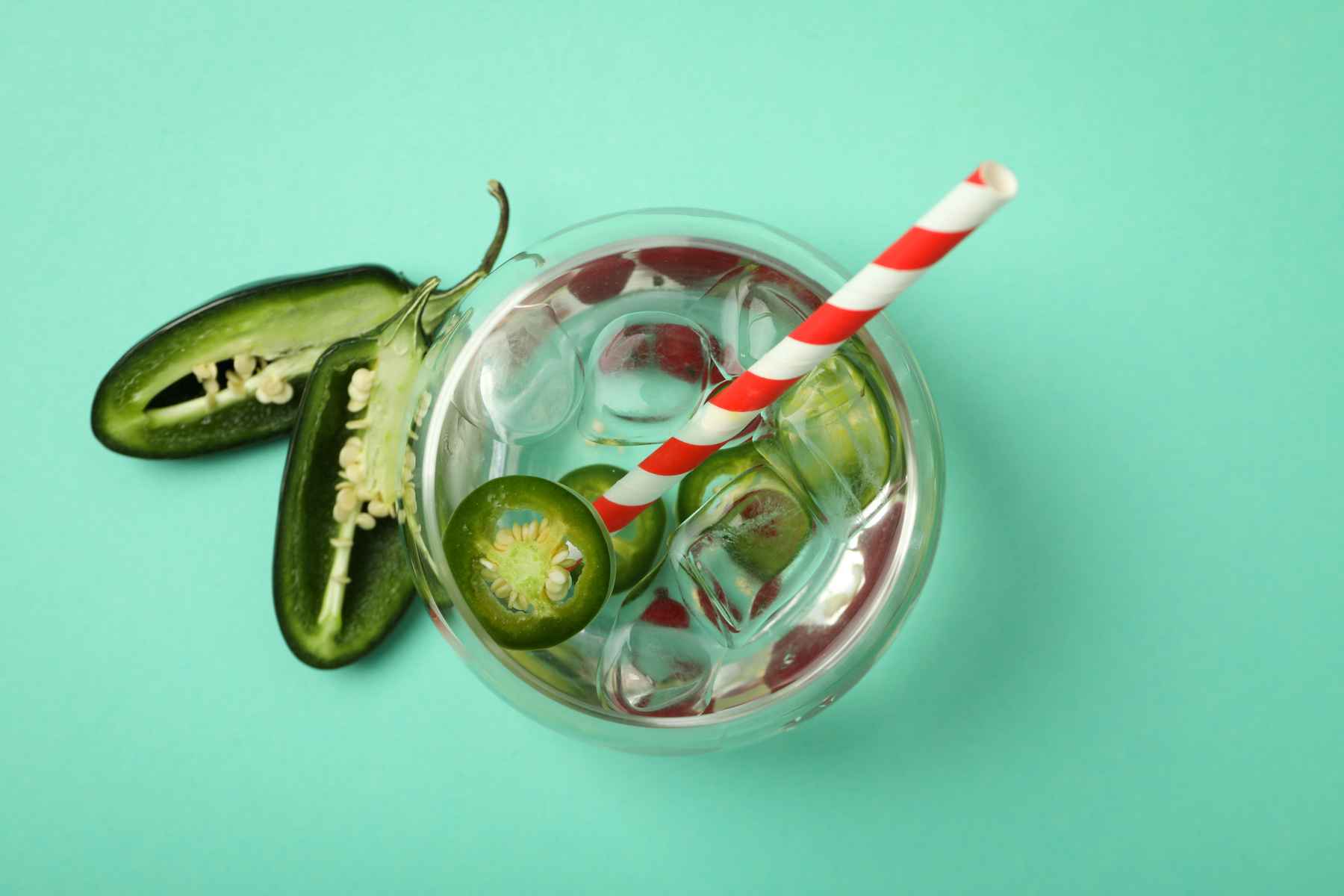 Overhead view of beverage garnished with halved jalapeno chiles with a red and white straw on teal background.