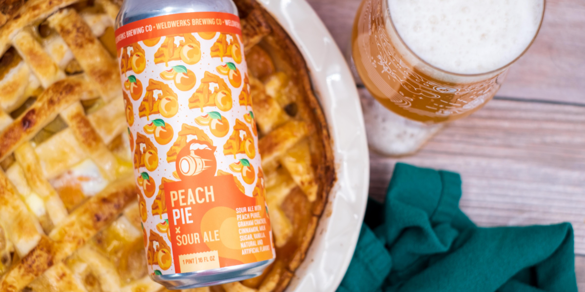 A can of peach pie beer by Weldworks Brewing sitting on a peach pie with a glass of beer next to it on a wood table