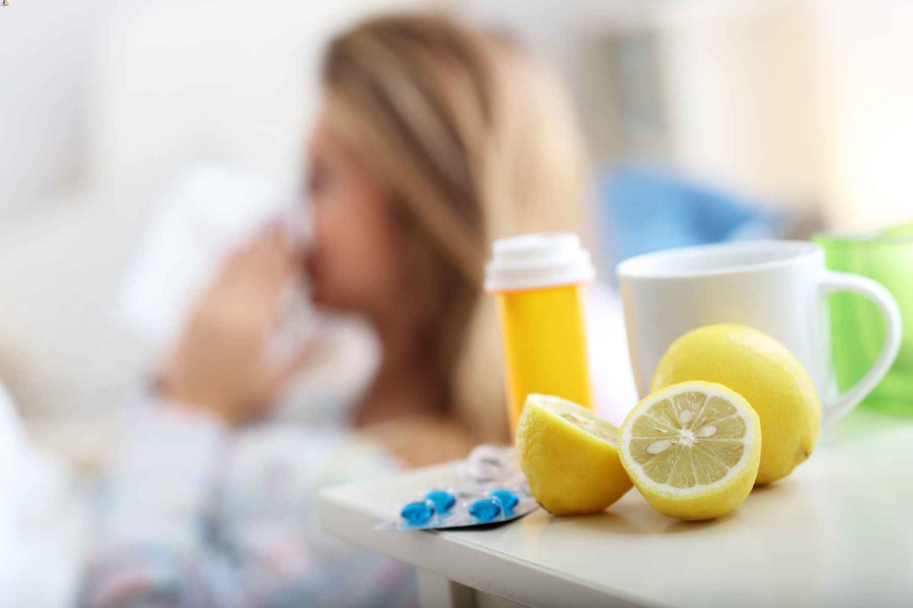 Out of focus woman in background blowing her nose, with cup of tea, lemons, and medication in focus in foreground.