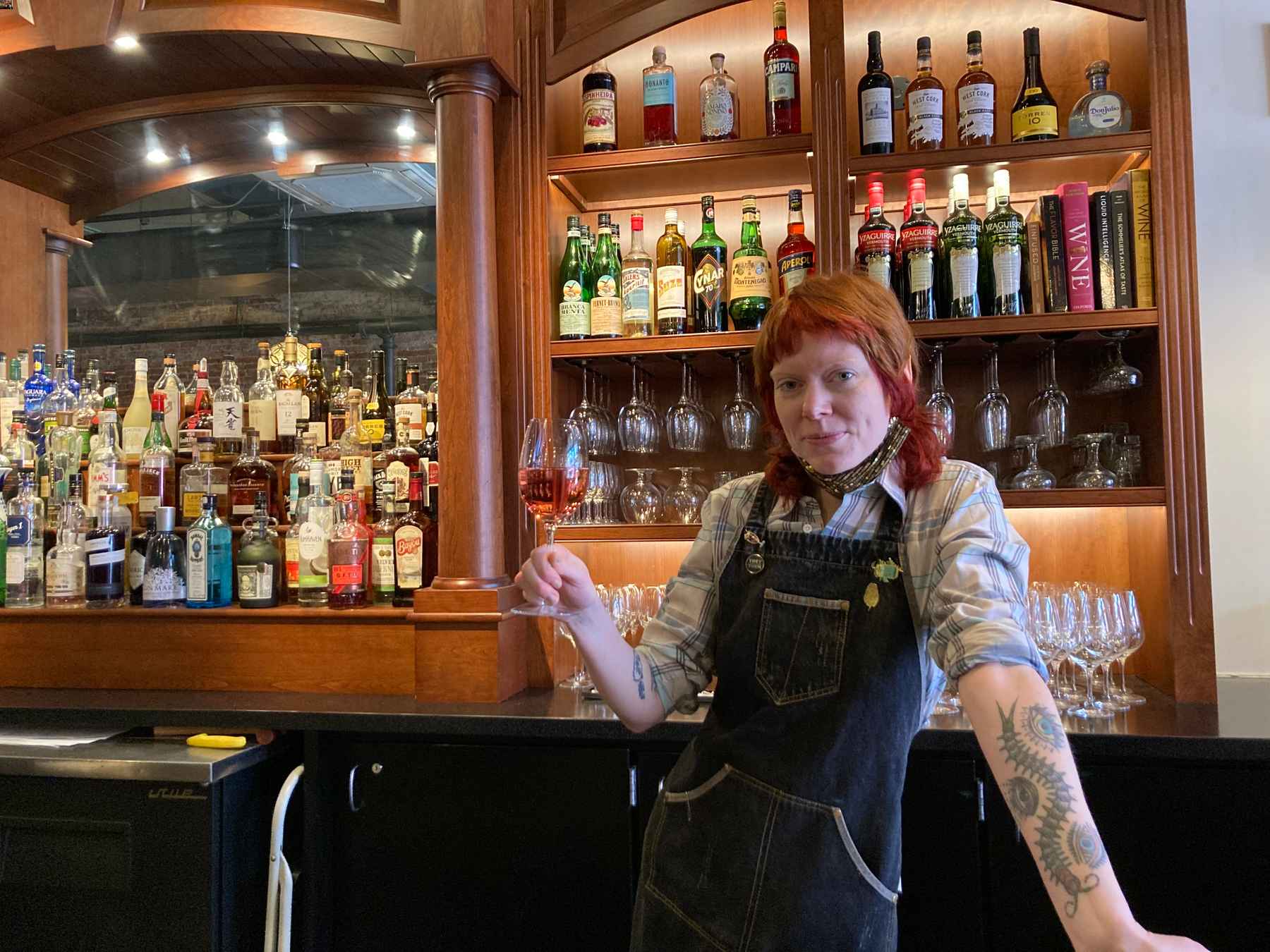 Bartender wearing apron with bright red hair and holding a glass of rose wine in front of the barback.