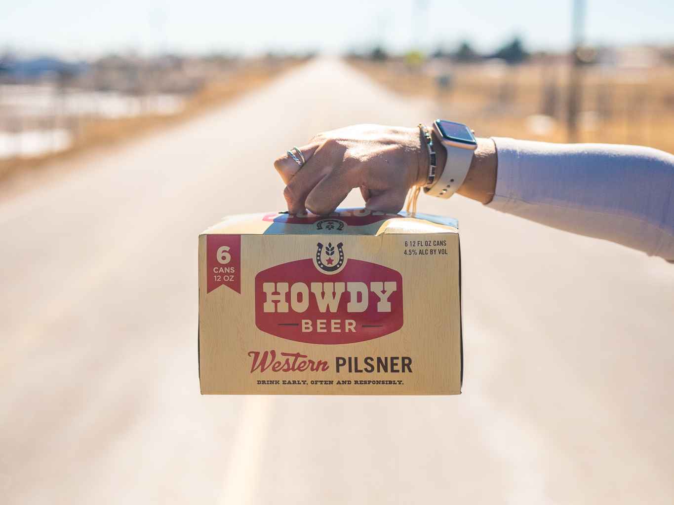 Hand holding a six-pack of Howdy Beer with an out of focus open road behind it.