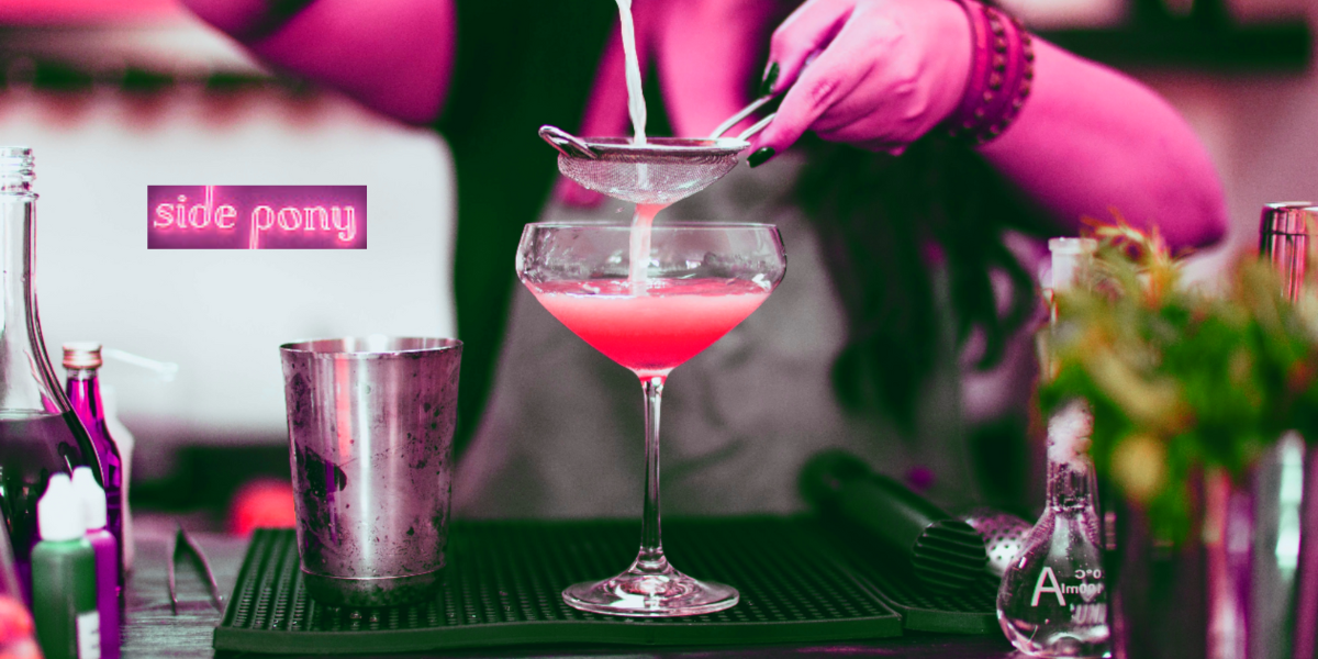 A bartender pours a drink through a strainer into a coupe glass in a photo that's black and white with pops of pink. The logo for the new Side Pony cafe & cocktail lounge in Denver appears on the upper left side.