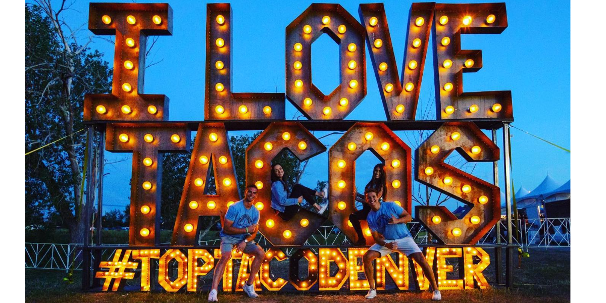 A giant sign of lit rustic letters spells out "I Love Tacos. Top Taco Denver" while people in blue shirts climb on the letters. Taken at Top Taco Festival, produced by DiningOut Events