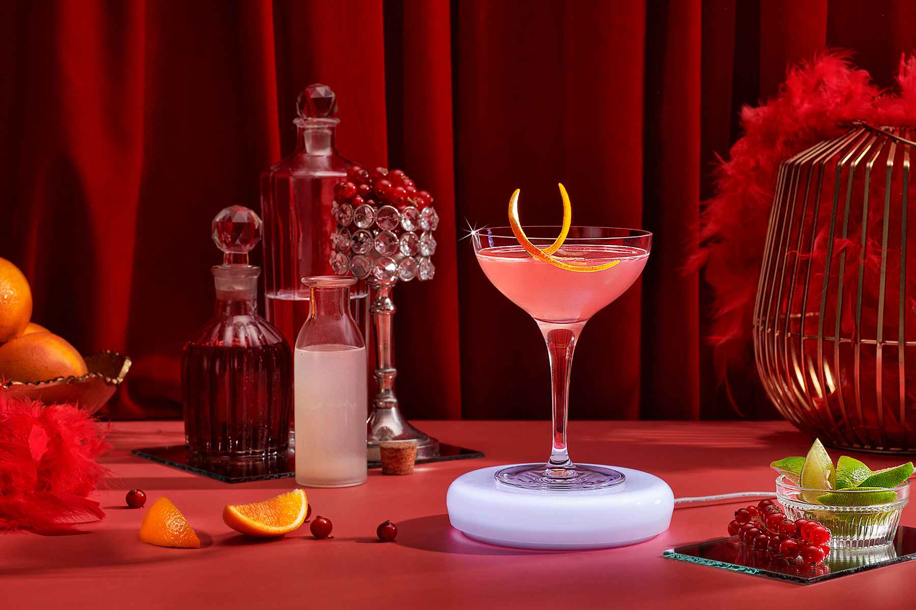 Pink cocktail in a coupe glass sitting on electronic Barsys coaster. Luxurious red background with velvet, feathers and liquor decanters.