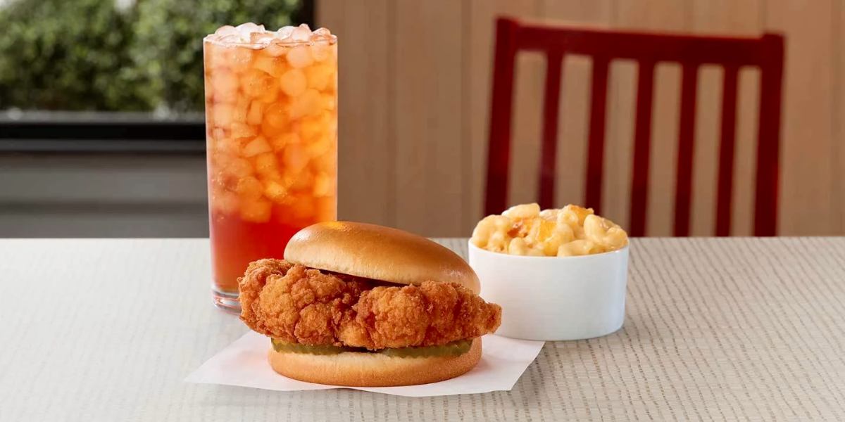 A plant-based "chicken" sandwich made from cauliflower on a table with a bowl of mac and cheese and a glass of iced tea with a red chair in the background Chick-fil-A Cauliflower Sandwich
