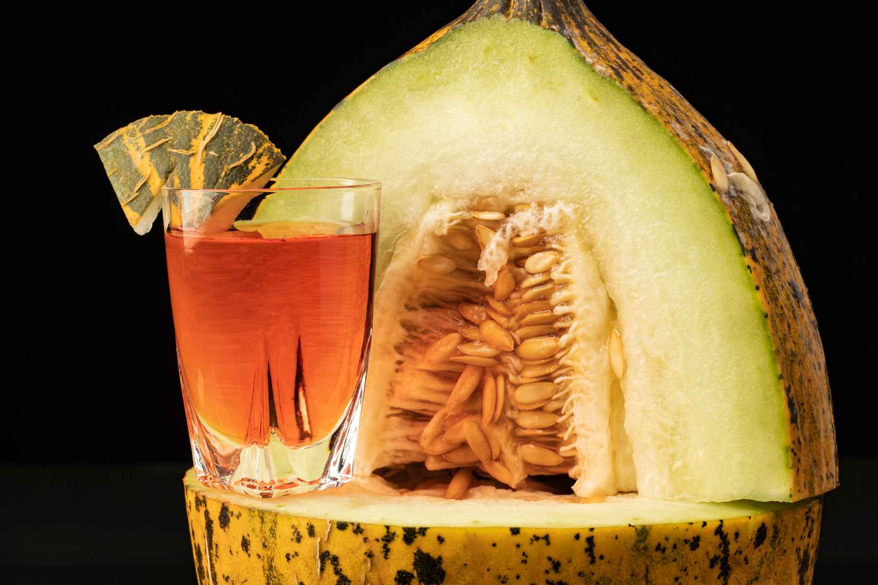 Glass of amber colored flavored spirits next to a melon half and garnished with a piece of melon peel.