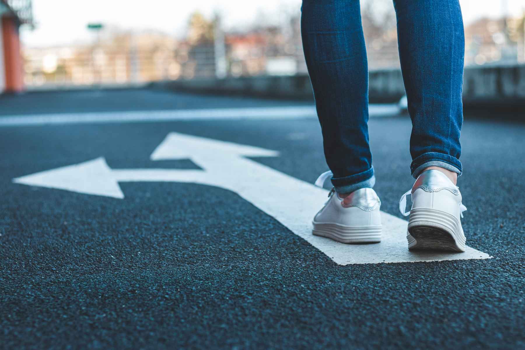 Woman wearing sneakers and walking down an asphalt road painted with straight and turn arrows. Staying or leaving.
