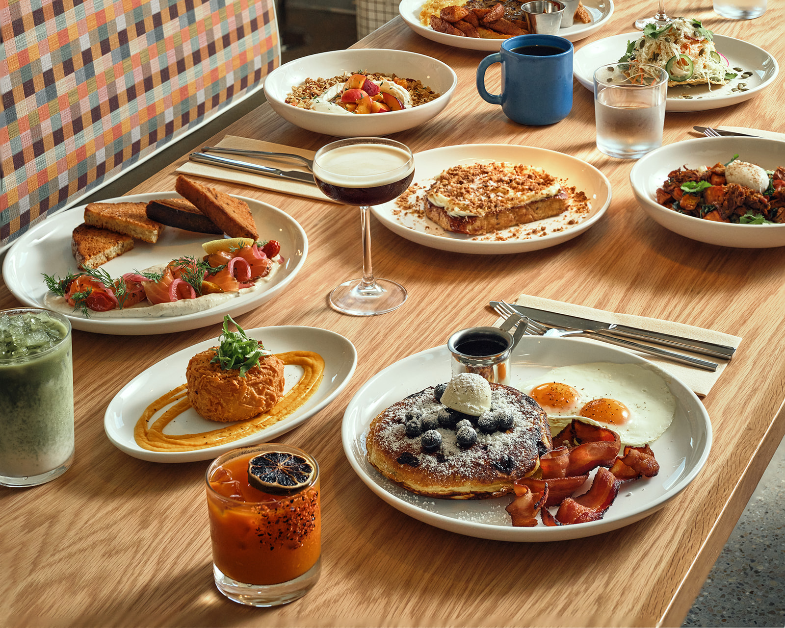 Brunch spread on a wooden table with multiple plates of salmon toast, smoked items, eggs, pancakes, yogurt bowls, a blue mug, and orange and green drinks.