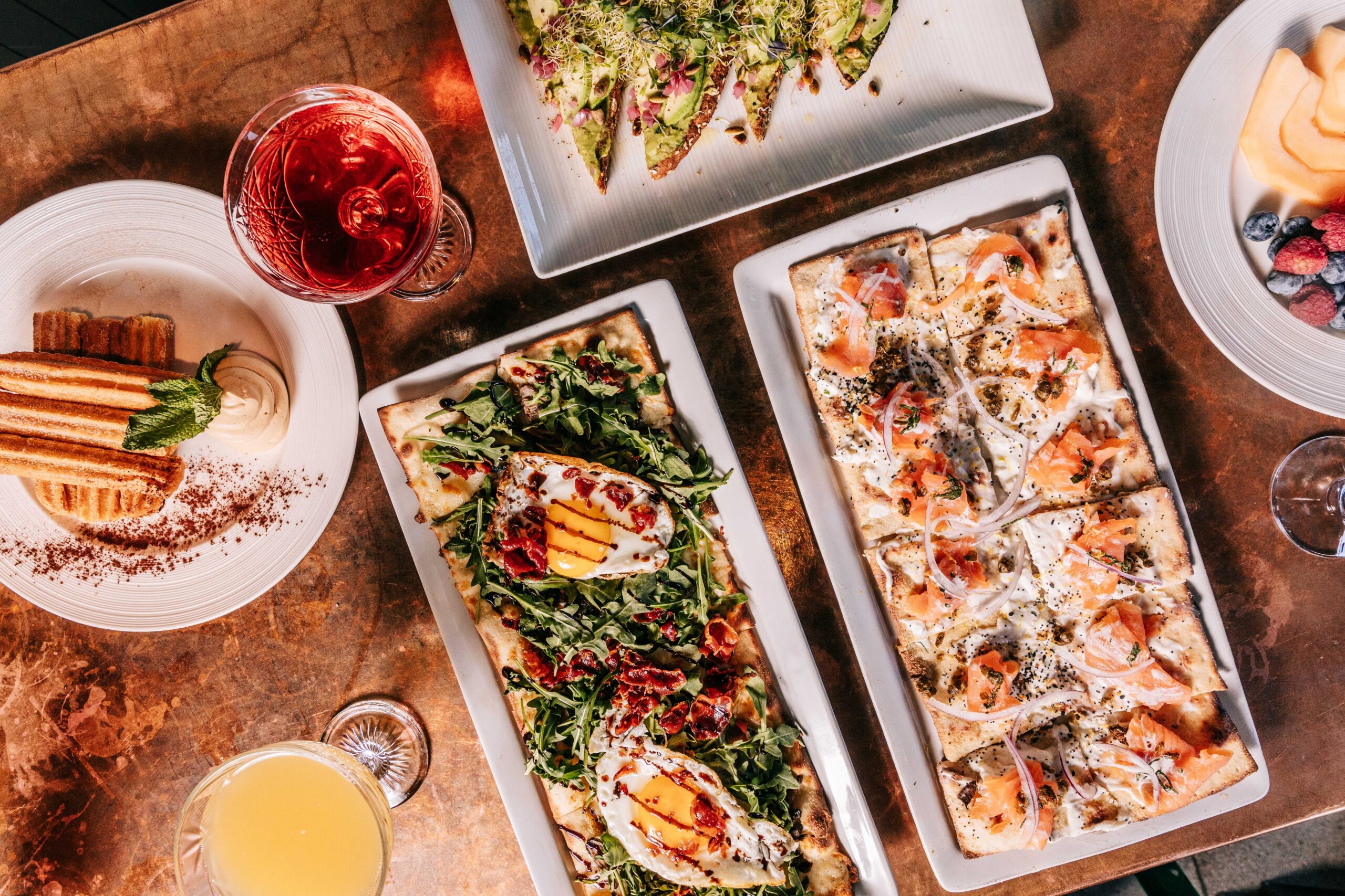 Top-down view of a spread: several rectangular flatbread pizzas with varied toppings, plus drinks and sides on a copper-toned table