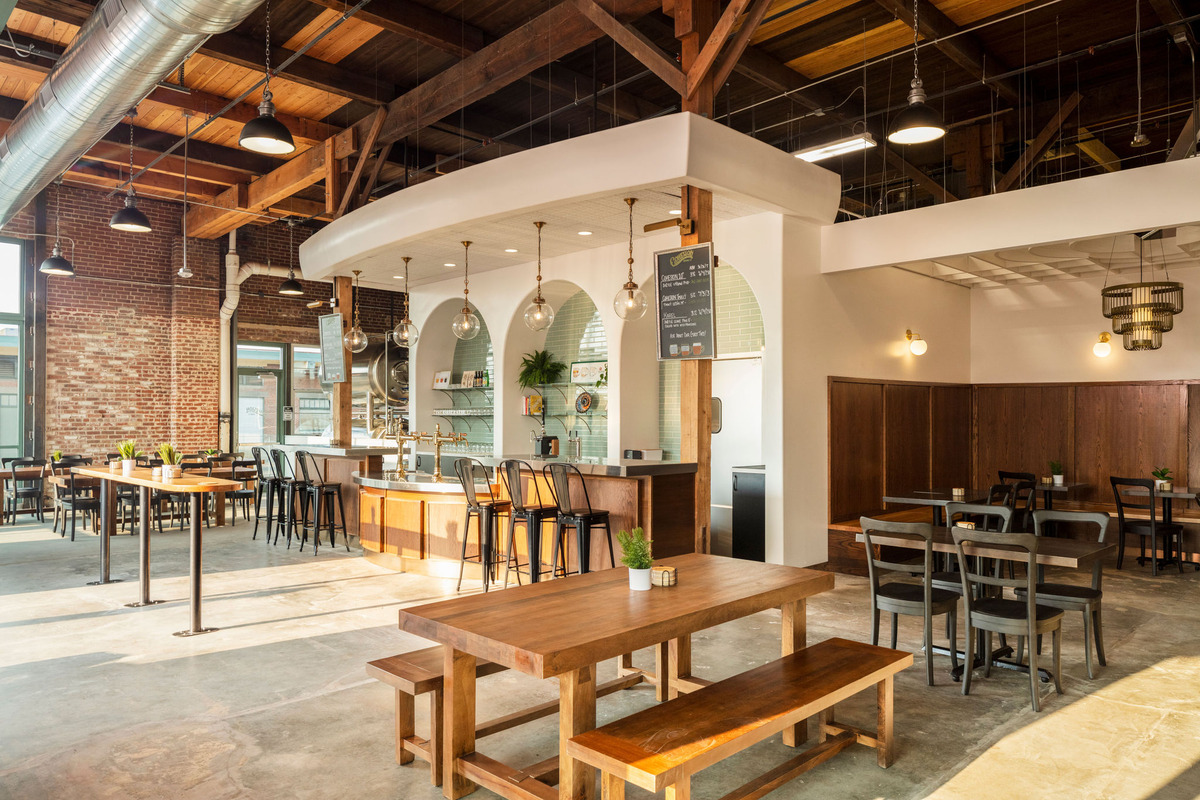 Bright industrial cafe interior with wooden beams, hanging glass lights, and a curved white counter with bar stools.