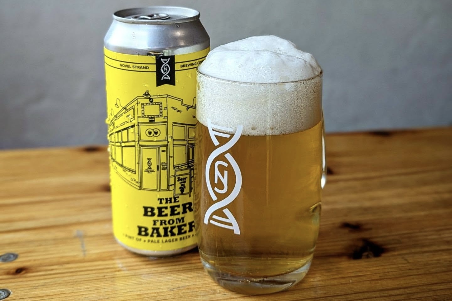 Yellow beer can with black line-art graphics beside a tall glass of pale ale on a wooden table, foam overflowing slightly at the rim.