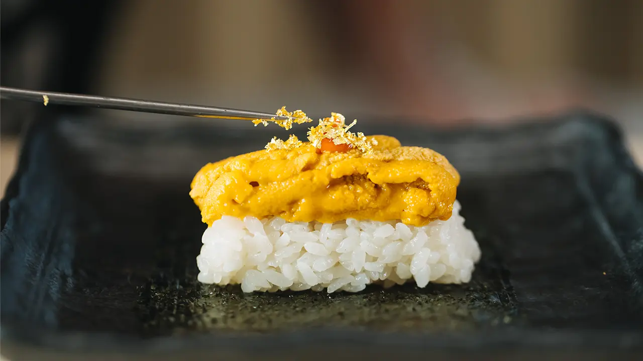 nori - DiningOut Nigiri sushi with white rice topped by sea urchin, with edible gold leaf being added by tweezers on a dark plate