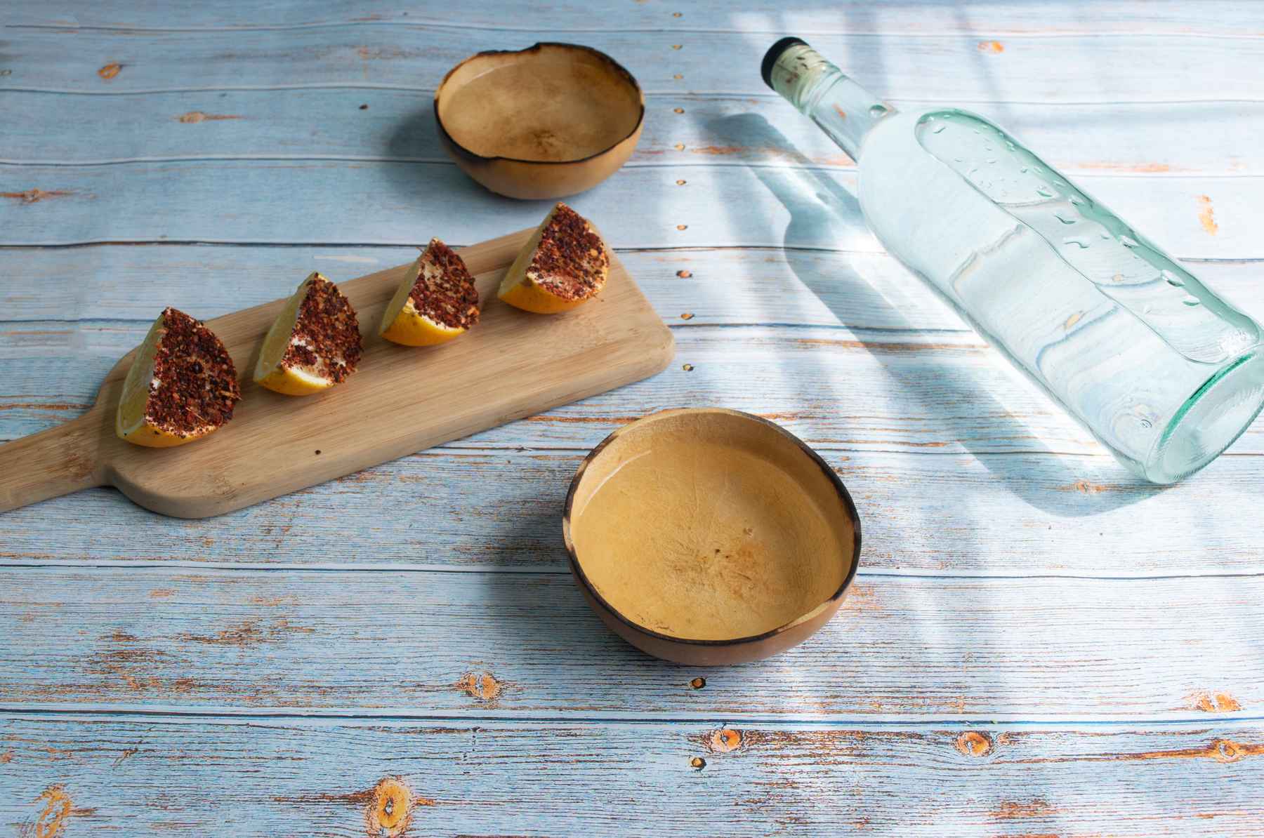 Bottle of mezcal next to orange slices dipped in red chile and two copitas full of mezcal.