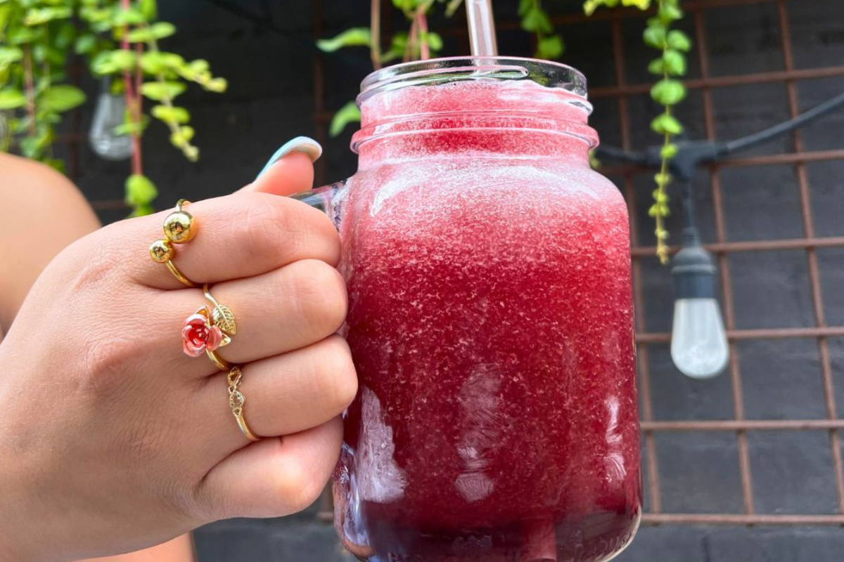 A close-up of Acova’s Misty, a frozen red cocktail served in a mason jar, held by a hand wearing gold rings with a plant-filled patio in the background.