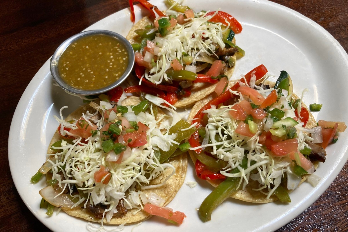 Three tacos from Adelitas topped with sautéed peppers, onions, shredded cabbage, and fresh pico de gallo, served with a side of green salsa on a white plate.