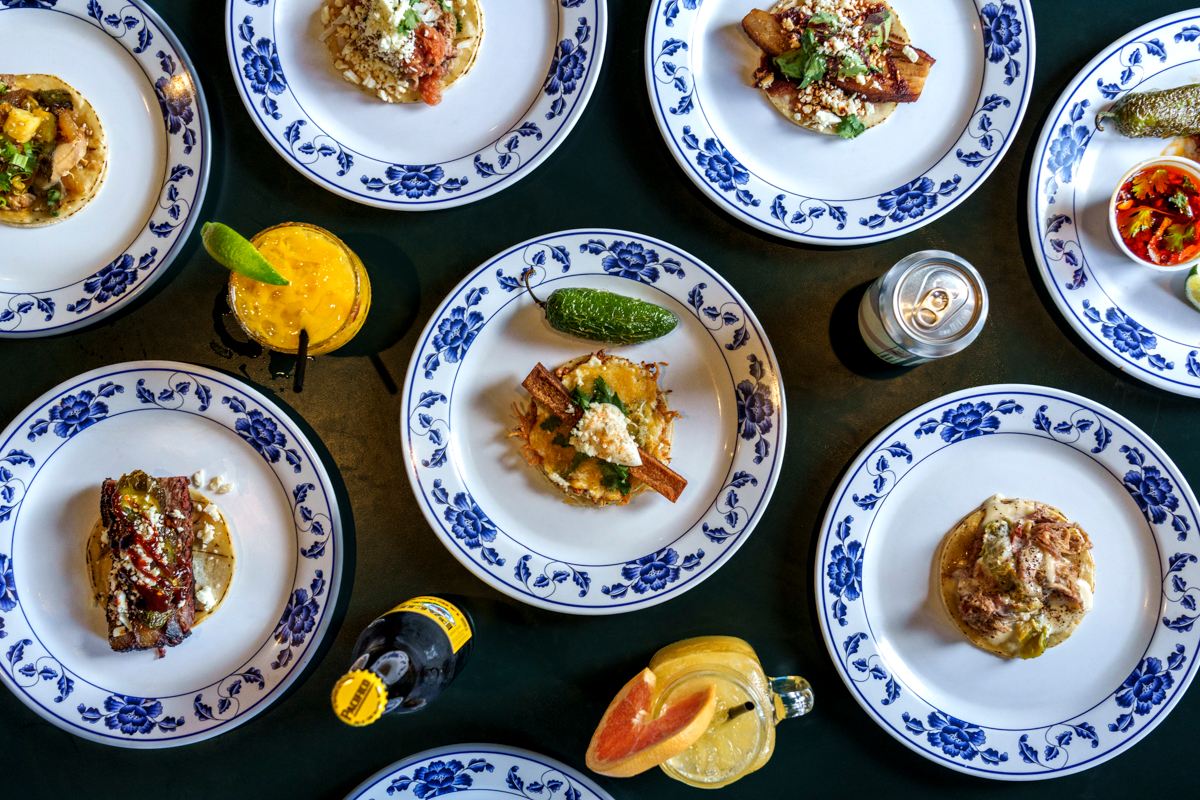 An overhead view of Adobo’s colorful taco spread, each plated on floral china, paired with cocktails, soda, and garnished with fresh lime and jalapeños.









