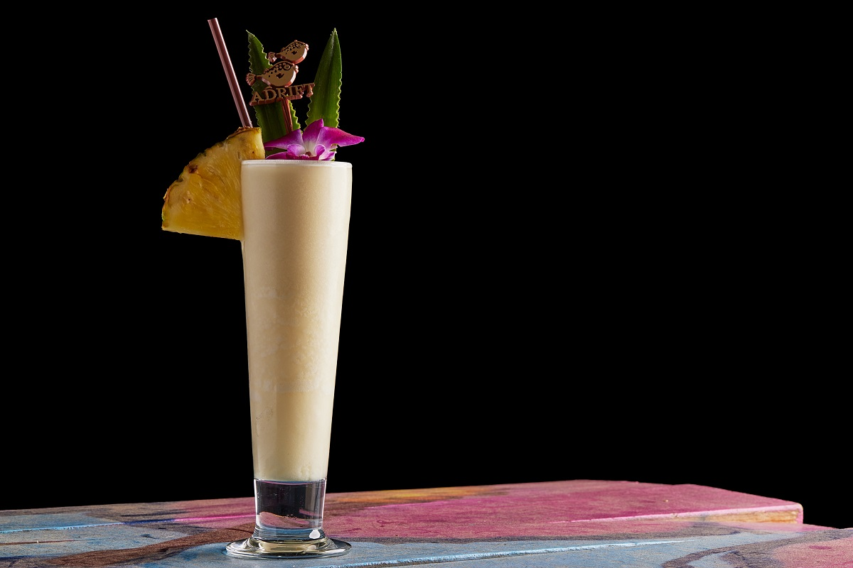 Adrift’s Chi Chi cocktail served in a tall glass with pineapple, orchid, and palm leaf garnish on a colorful wood table with a black background.