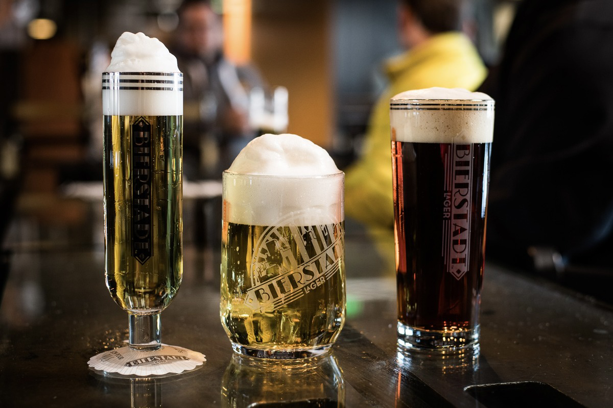 Three glasses of Bierstadt lager with thick foam heads, showcasing light and dark varieties on a reflective bar counter at a brewery or bar.