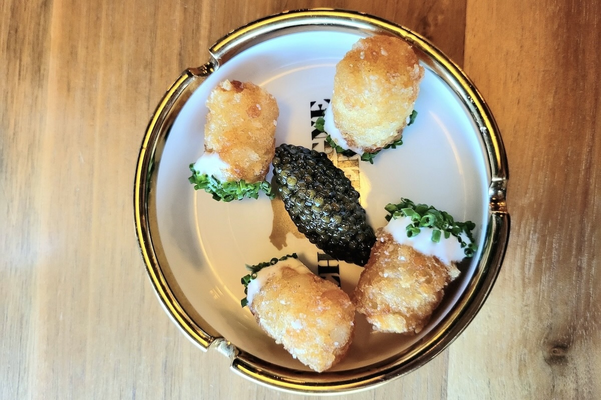 A luxe ashtray-style dish holds five golden tater tots with sour cream and chives, arranged around a generous spoonful of black caviar in the center.