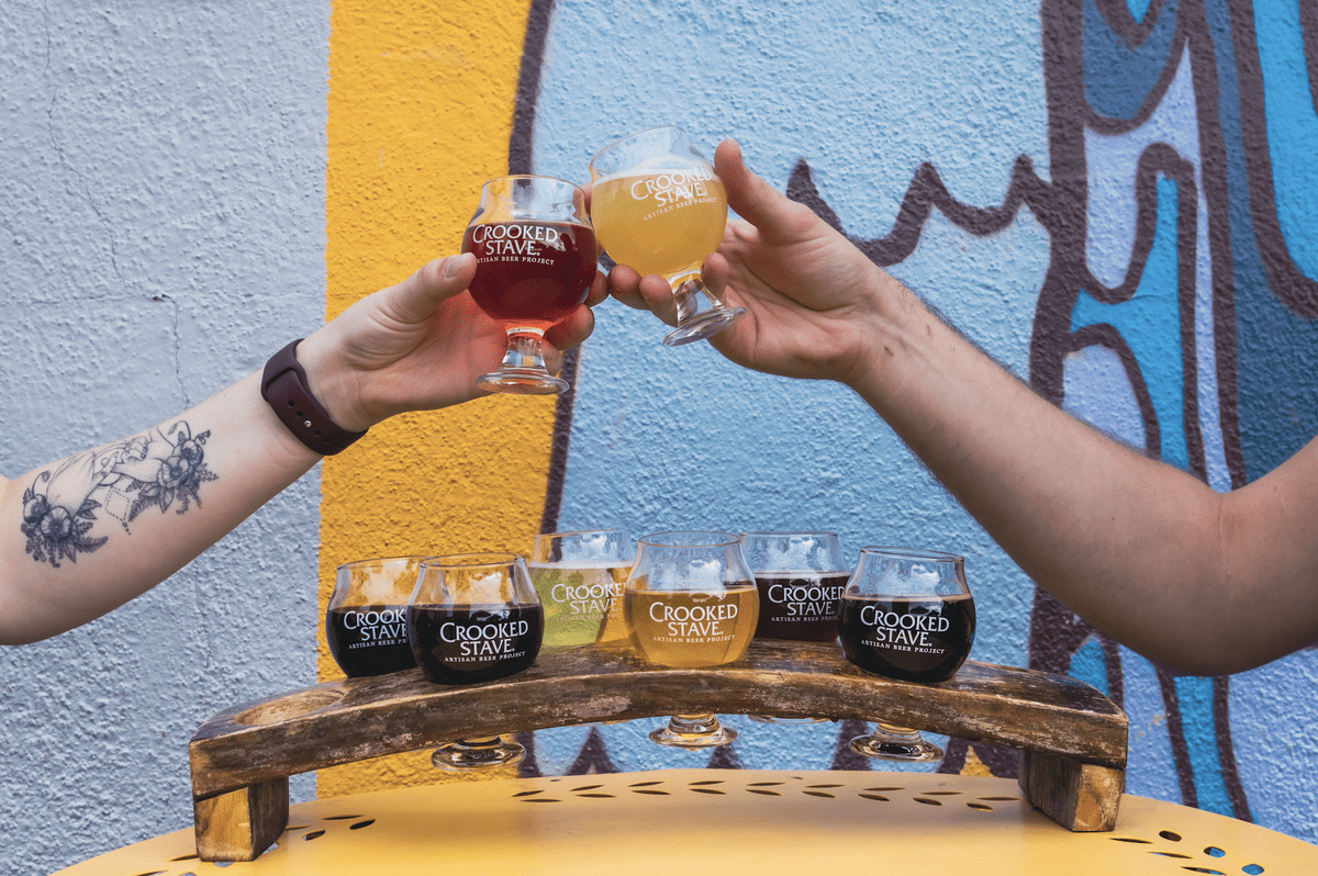 Two people cheers with Crooked Stave beers over a colorful wall, with a rustic beer flight tray featuring various artisan brews on a yellow table.