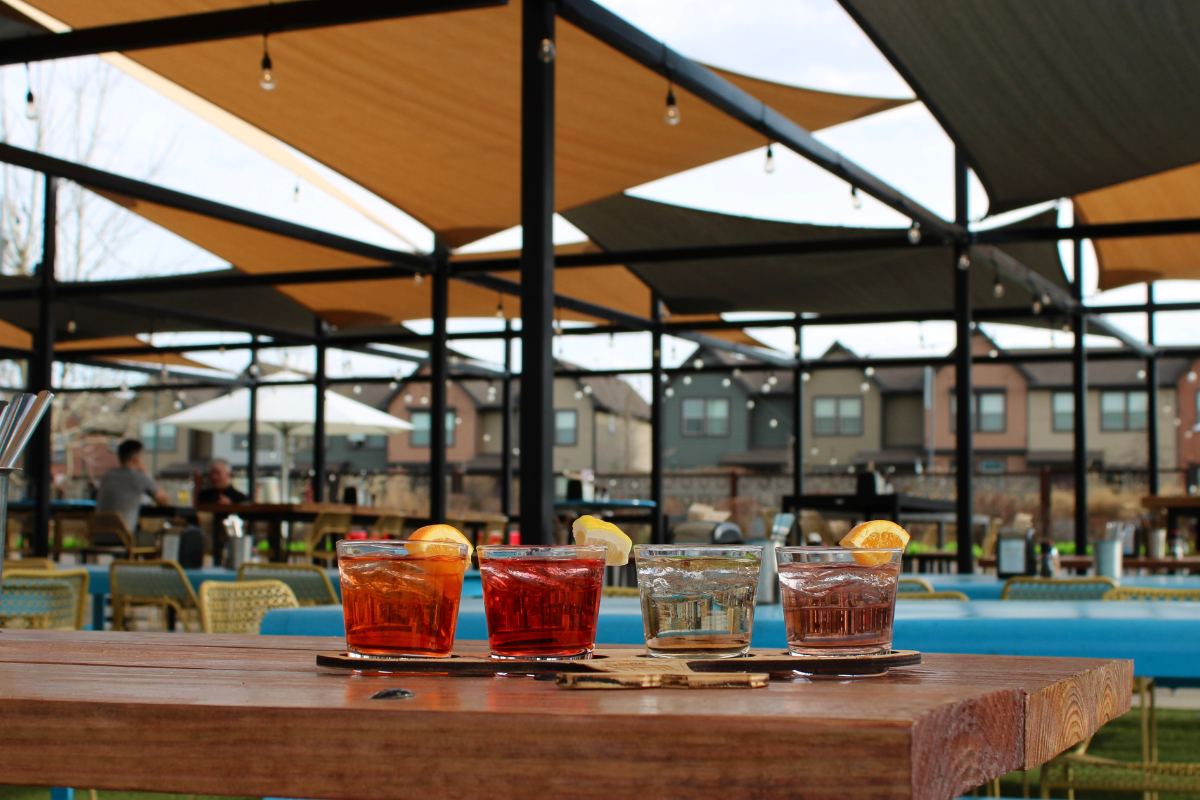 12 Must-Visit Restaurants in Bishop Arts District - DiningOut Spritz flight with four colorful cocktails garnished with citrus, served on a wood table at FlyteCo Tower’s outdoor patio with shaded seating.