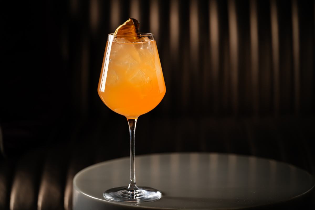 BishopArts_Pillar_PhotobyAdriana-Herrera - DiningOut Sunset Spritz cocktail with rum, Aperol, and pineapple shrub, served over ice in a wine glass on a round table at Halo rooftop bar in Denver.