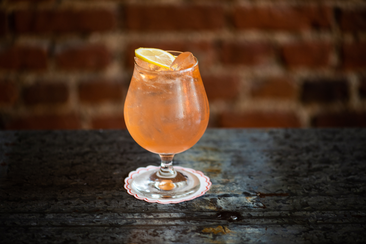 BishopArts_PotpourriBoulangerie_CourtesyofPotpourrioulangerie - DiningOut Casoni Spritz served over ice in a stemmed glass with a lemon slice at Jovanina’s, placed on a metal table with a brick wall in the background.