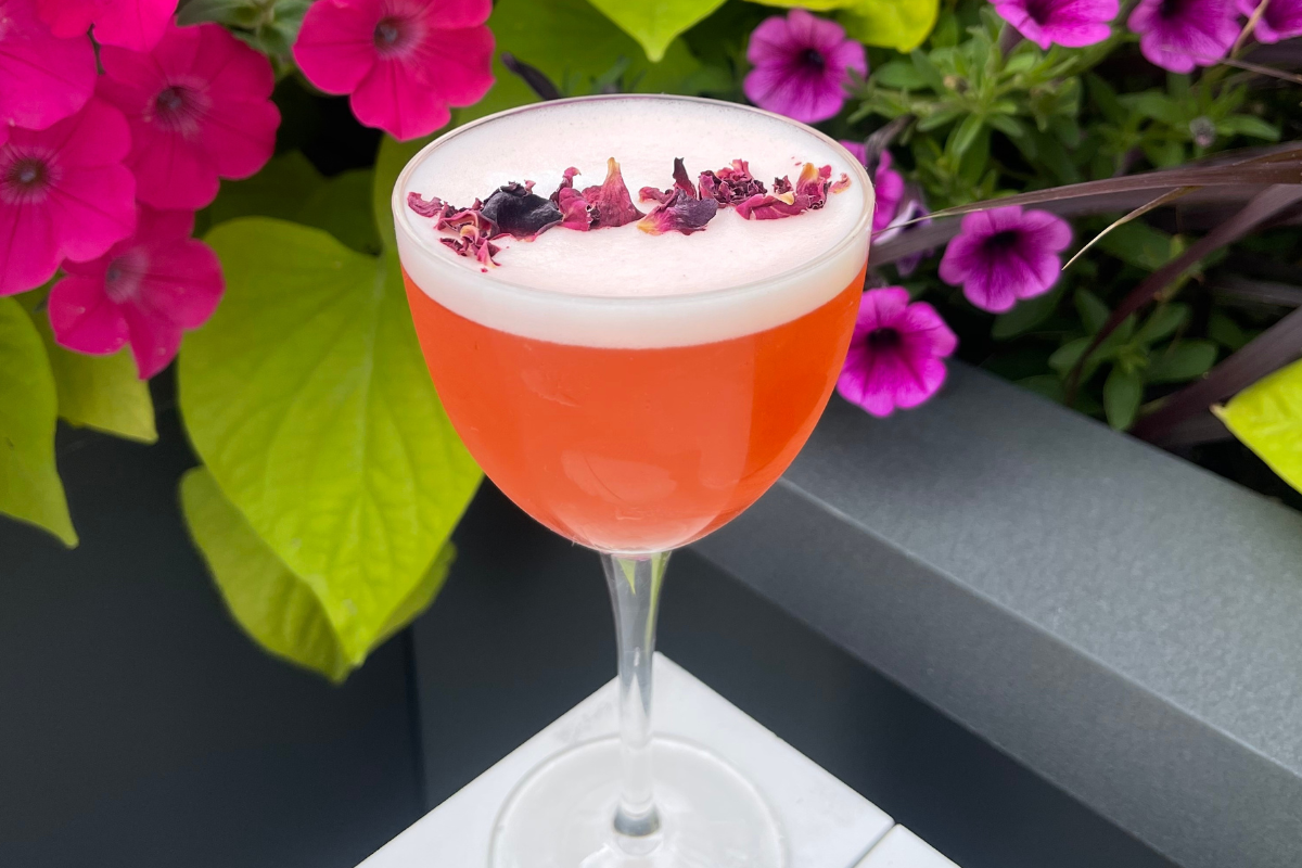 BishopArts_Pillarburger_PhotobyAdriana-Herrera - DiningOut Strawberry Tides spritz with a foamy top and dried flower garnish, served in a coupe glass at Kisbee on the Roof with bright flowers in the background.