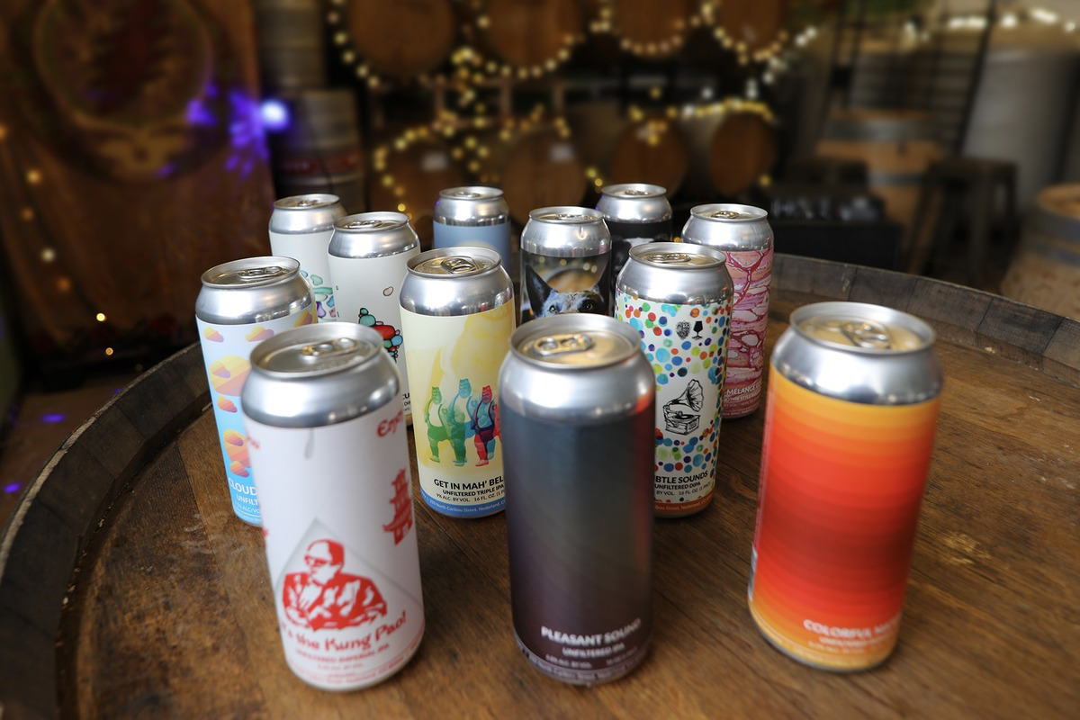 Colorful craft beer cans from Knotted Root Brewing Company displayed on a barrel top, with festive string lights and barrels in the background.