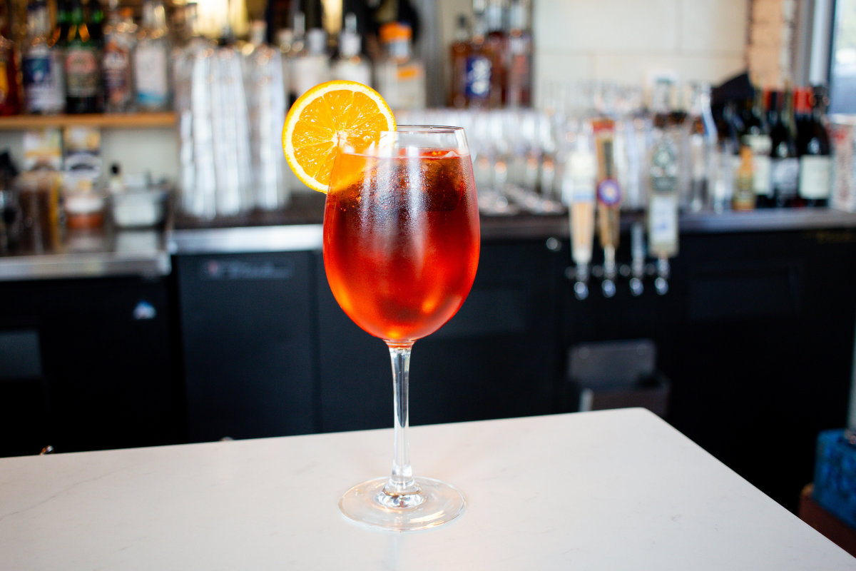 How Grief Shaped Georgie's Chef RJ Yoakum Into One of the Most Ambitious Chefs in Dallas - DiningOut Leven Deli Co.’s house spritz served in a wine glass with ice and an orange slice garnish, set on a white counter with a bar backdrop.