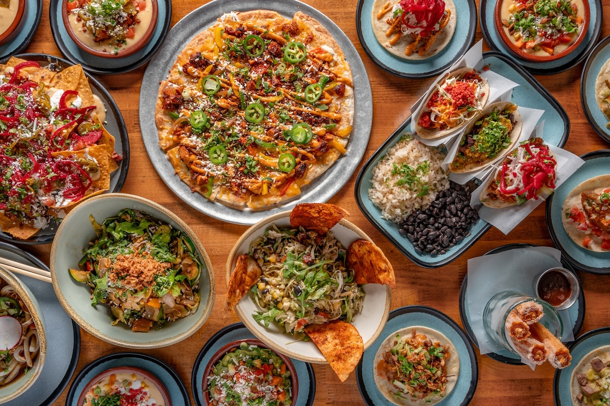 Colorful spread from Los Chingones featuring tacos, nachos, flatbread, rice and beans, salads, queso dips, and churros served on a wood table.









