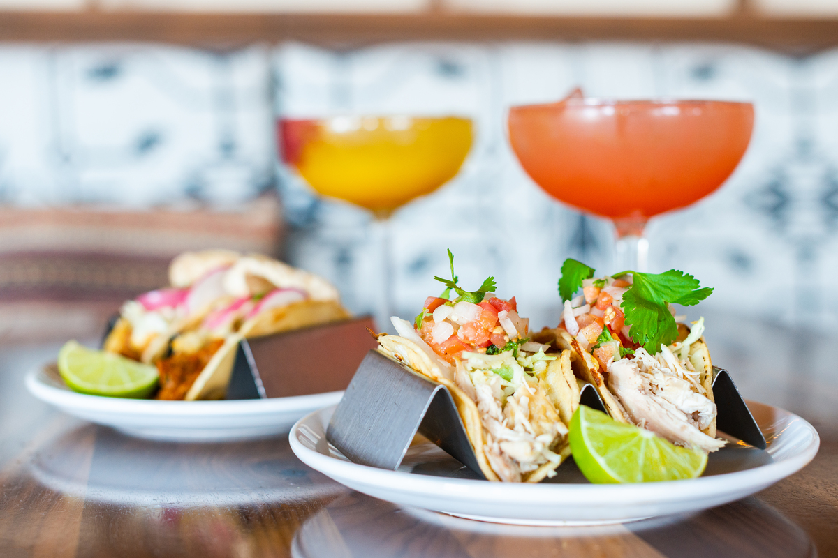 Two plates of tacos from My Neighbor Felix are served with lime wedges and topped with shredded chicken, pico de gallo, and cilantro, with cocktails in the background.