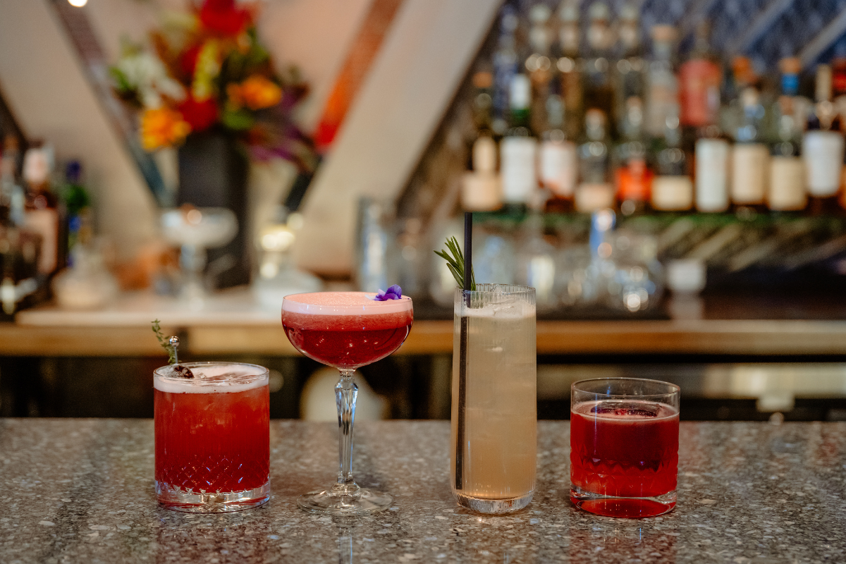 Georgie-Dining-Room_Nicole-Frazen-1 - DiningOut Four colorful zero-proof spritz cocktails garnished with herbs and flowers, lined up on a marble bar counter at Nocturne jazz and supper club in Denver.