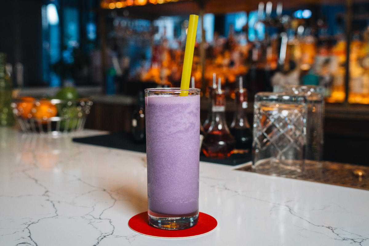 A tall glass of Poka Lola Social Club’s Bad News Bears slushy cocktail, bright purple in color with a yellow straw, served on a white marble bar.