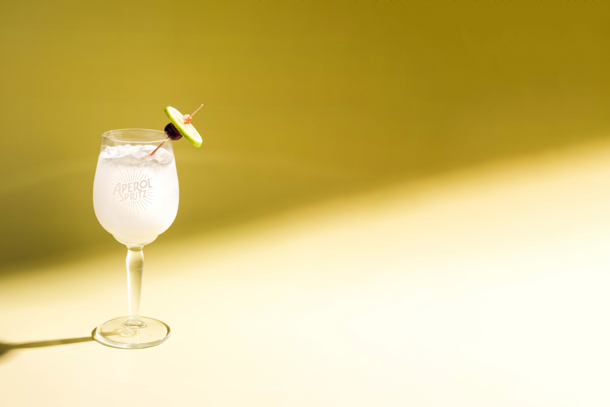 Georgie_Halibut_CourtesyofTravisStreetHospitality - DiningOut Coco Blanco Spritz from Punch Bowl Social, a light coconut cocktail served over ice in an Aperol glass with a lime and cherry garnish on a yellow backdrop.