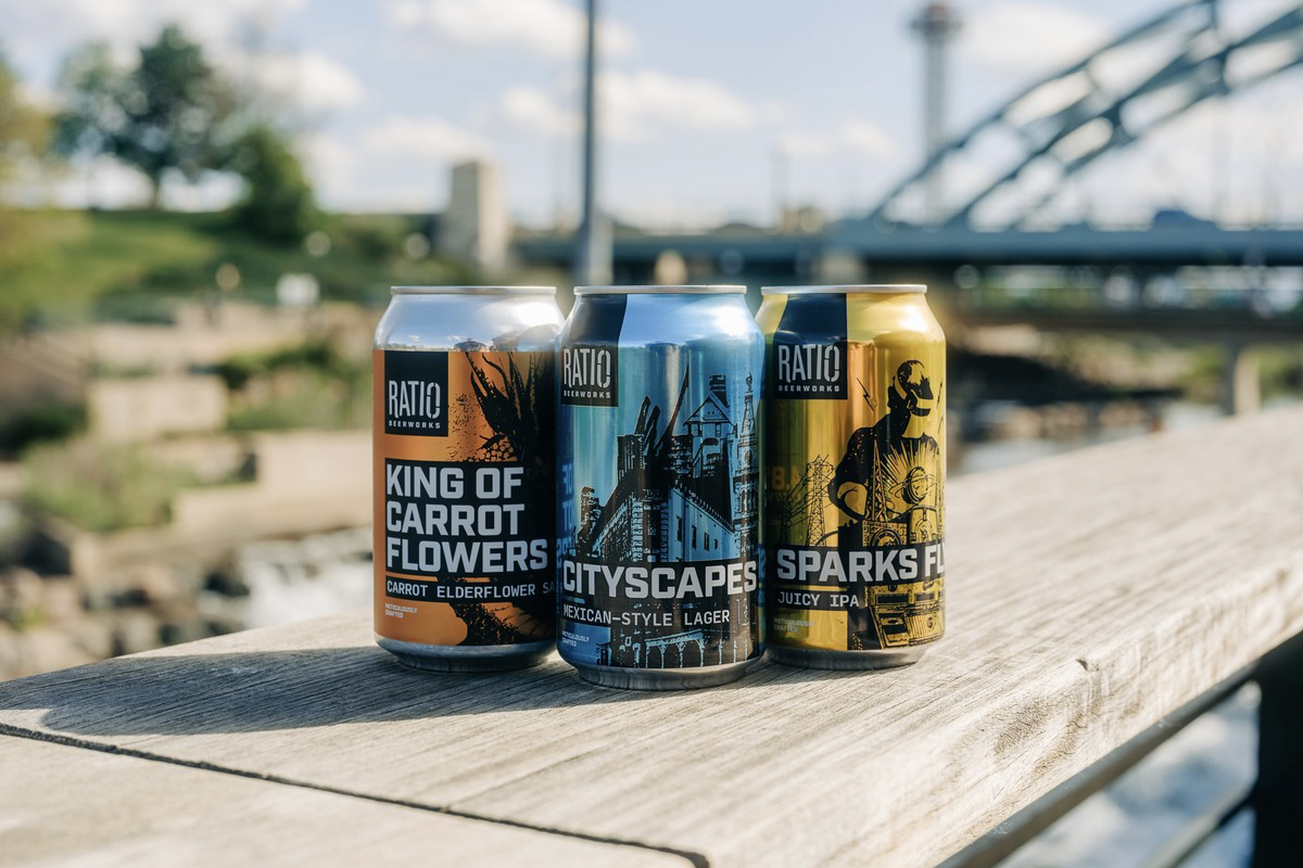 Three Ratio Beerworks cans—King of Carrot Flowers, Cityscapes, and Sparks Fly—displayed outdoors on wood railing with bridge and skyline backdrop.