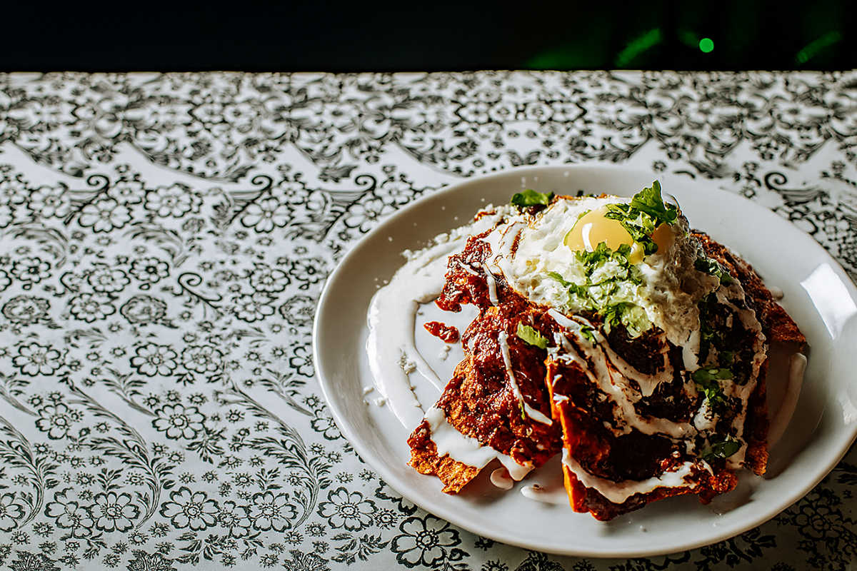 A plate of red mole chilaquiles topped with a runny fried egg, crema, crumbled cheese, and fresh cilantro sits on a white plate over a black-and-white floral tablecloth, lit from the side with soft, warm light.