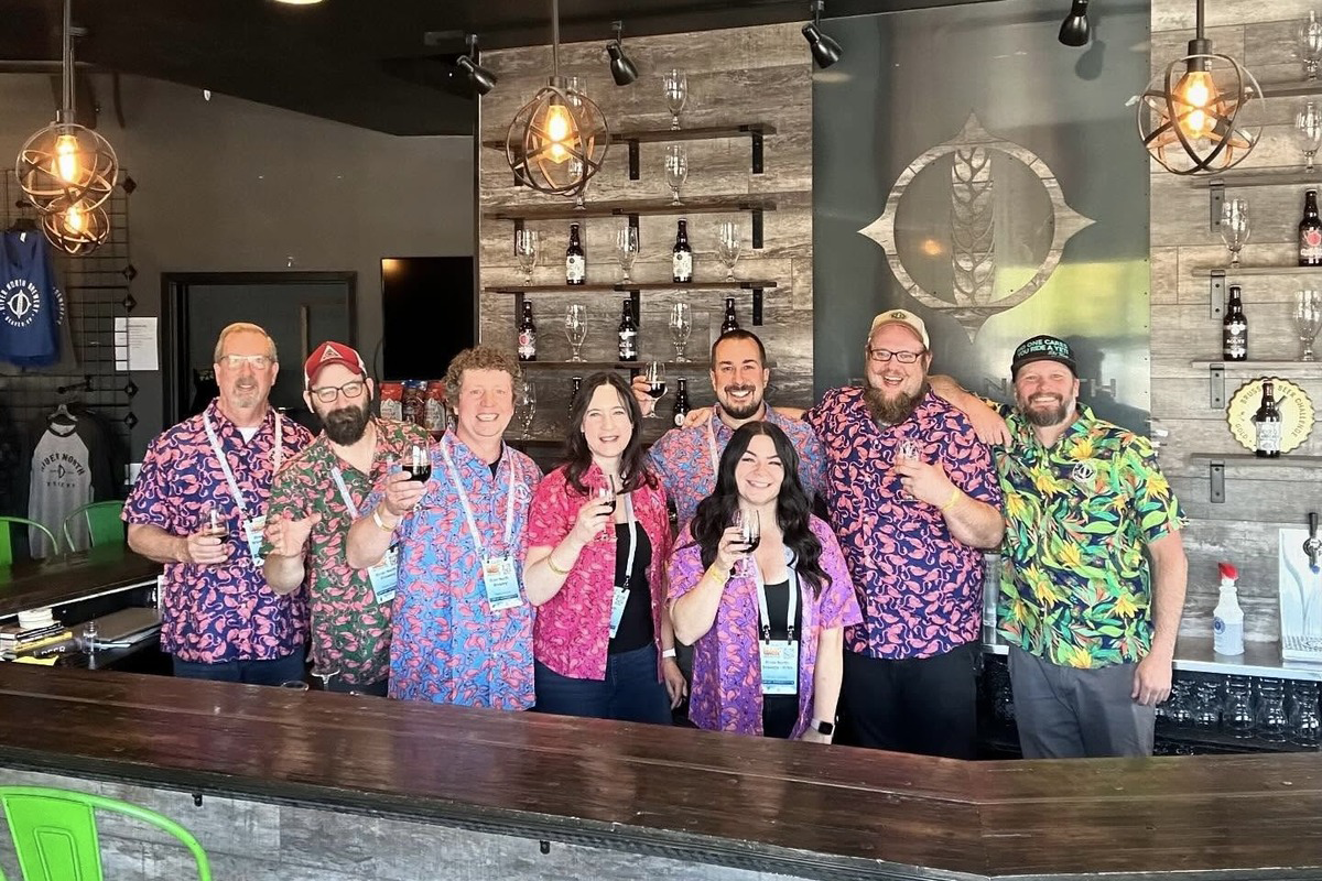 River North Brewery staff and brewers in colorful shirts smiling behind the bar, holding beer samples during a brewery event or beer festival in Denver.